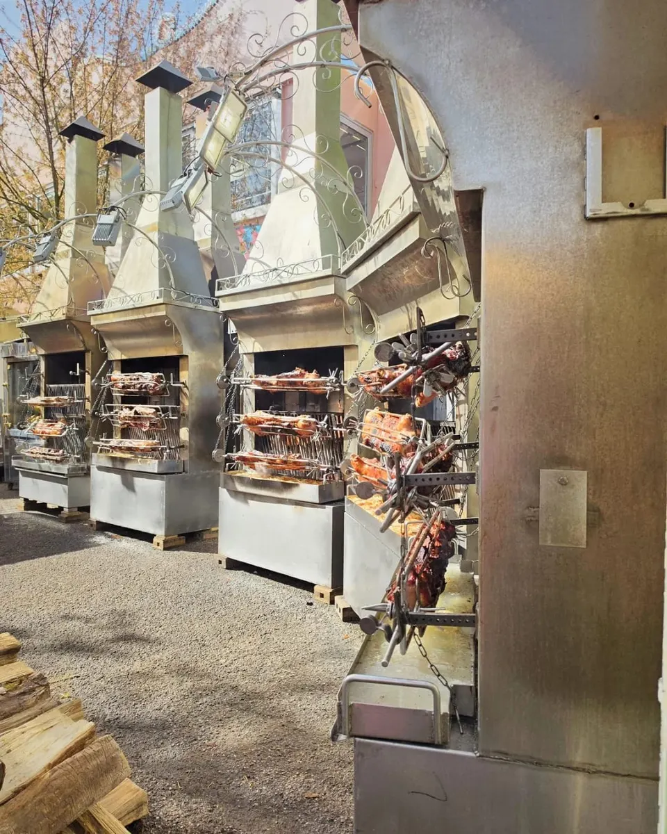 Alignement de rôtissoires en inox où des cochons et des pièces de viande cuisent embrochés au-dessus des braises, avec des bûches empilées au premier plan