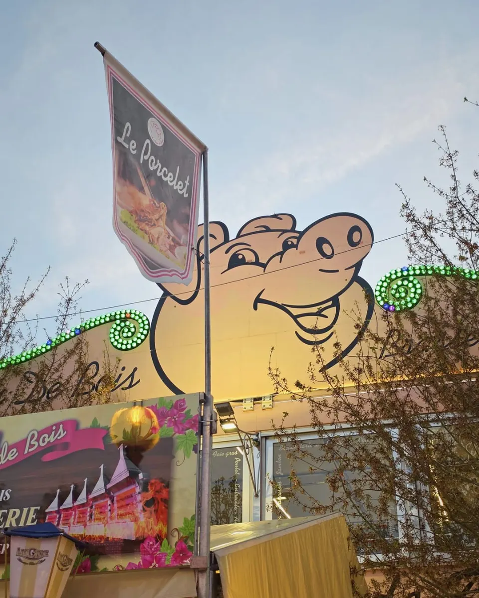 Vue en contre-plongée de la mascotte cochon géante posée sur le toit du restaurant foraine, à côté d'une oriflamme Le Porcelet, dans un ciel de fin de journée