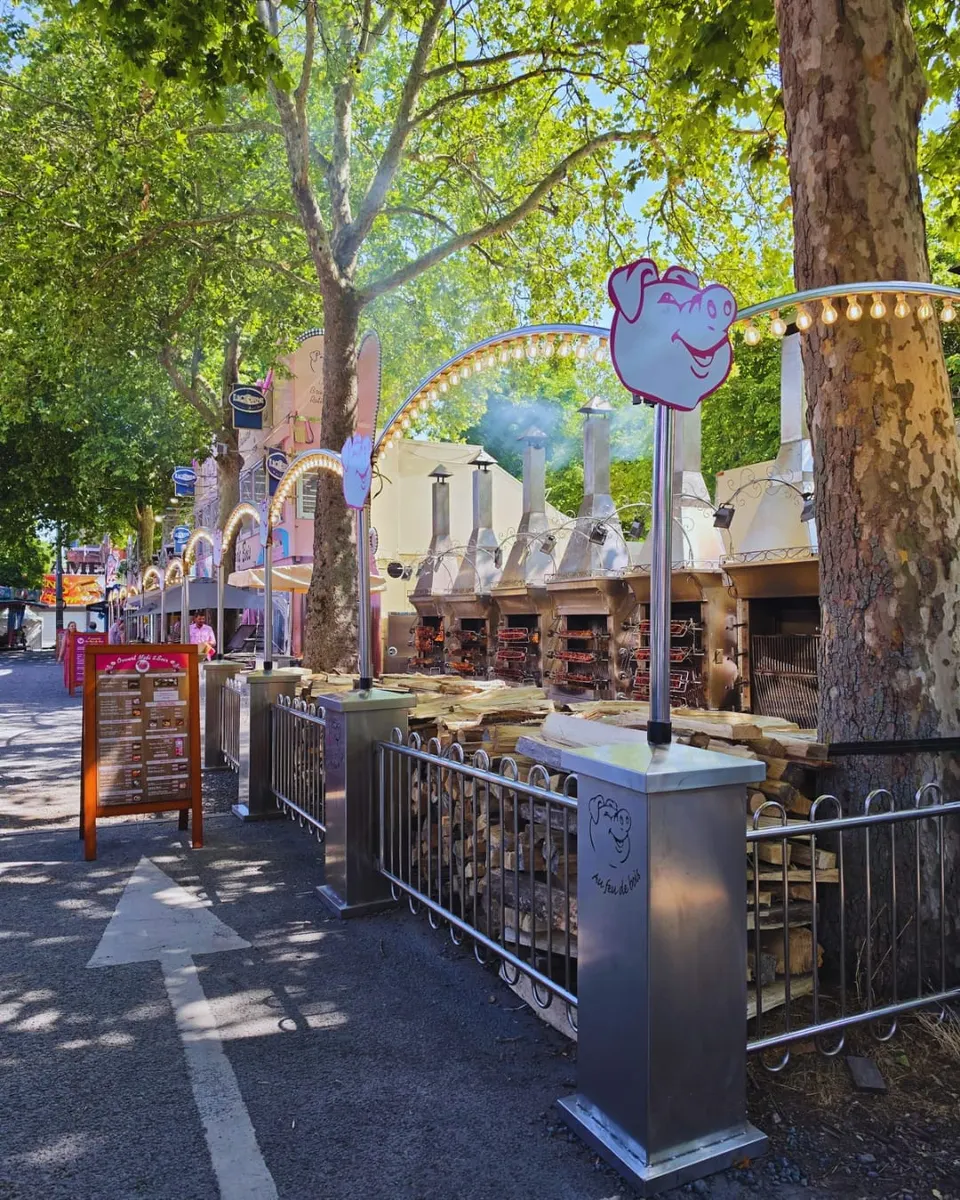 Allée ombragée bordée de platanes longeant la rôtisserie, avec des broches de viande qui cuisent devant un grand tas de bois fendu et des arches lumineuses décorées d'une tête de cochon.
