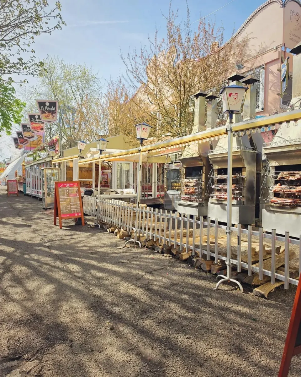 Allée ensoleillée de la foire longeant la façade du restaurant, avec des oriflammes, des guirlandes lumineuses et les rôtissoires en inox garnies de viandes