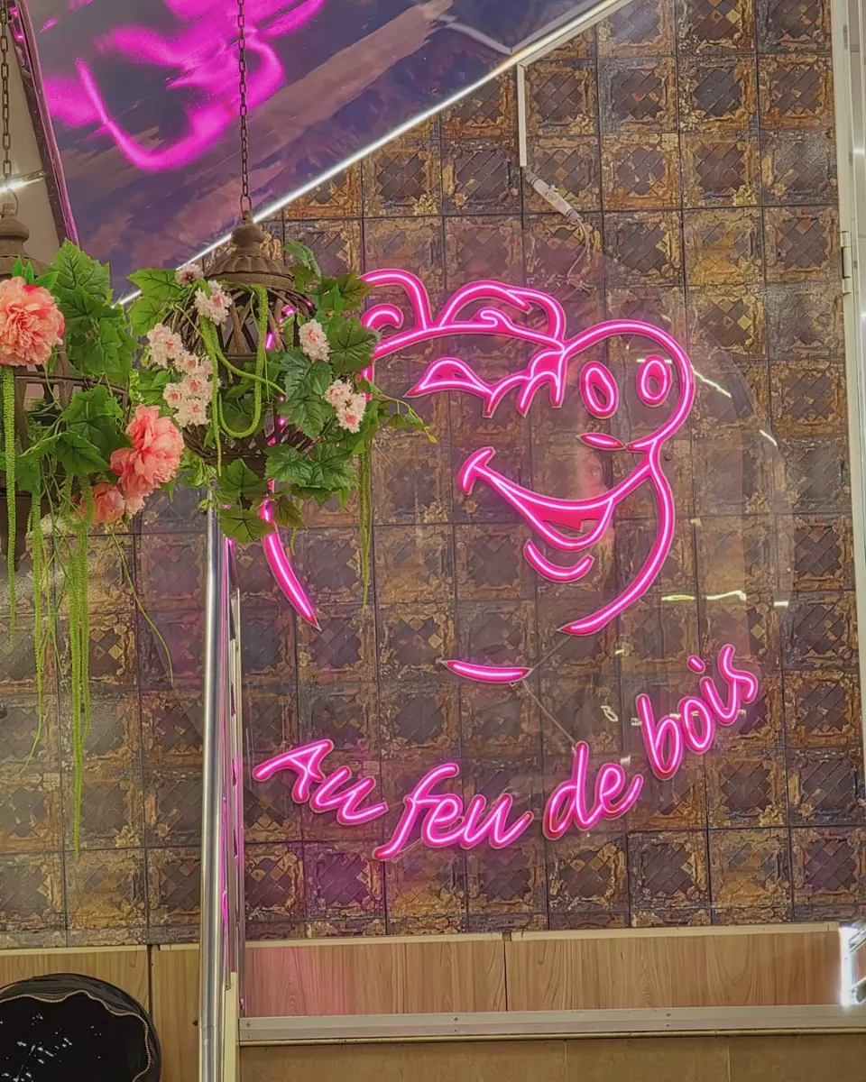 Enseigne au néon rose en forme de tête de cochon souriante avec la mention Au feu de bois, accrochée sur un mur de carreaux métalliques patinés, à côté d'une suspension de fleurs artificielles roses.