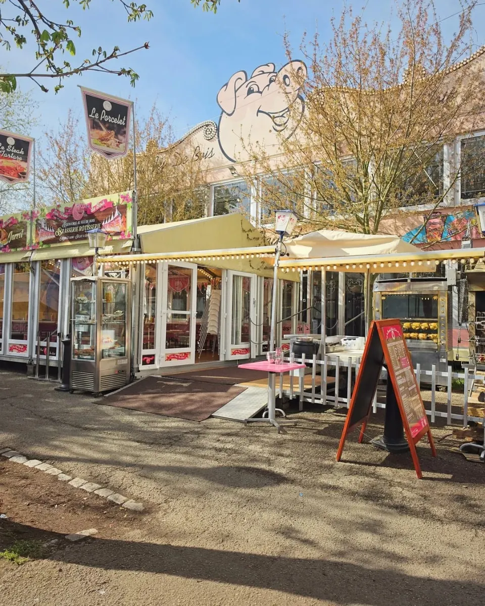 Façade ensoleillée du restaurant foraine avec la mascotte cochon sur le toit, une vitrine réfrigérée, une table haute rose et un chevalet de menu