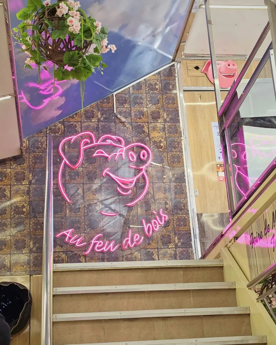 Enseigne néon rose en forme de tête de cochon portant la mention Au feu de bois, installée au dessus d'un escalier intérieur carrelé
