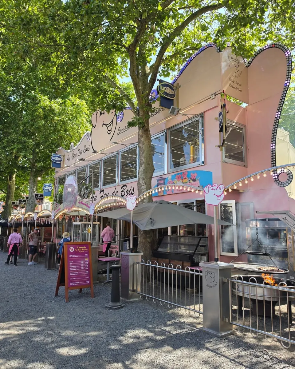 La façade rose de la brasserie, sa mascotte cochon et ses guirlandes d'ampoules, avec le grand grill au feu de bois qui brûle devant