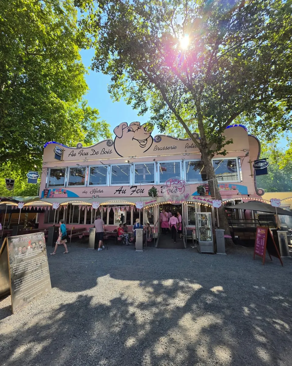 Le restaurant foragin rose Au Feu De Bois vu de face au soleil, avec l'équipe en chemises vichy roses devant l'entrée et des visiteurs qui passent sur l'allée de gravier.