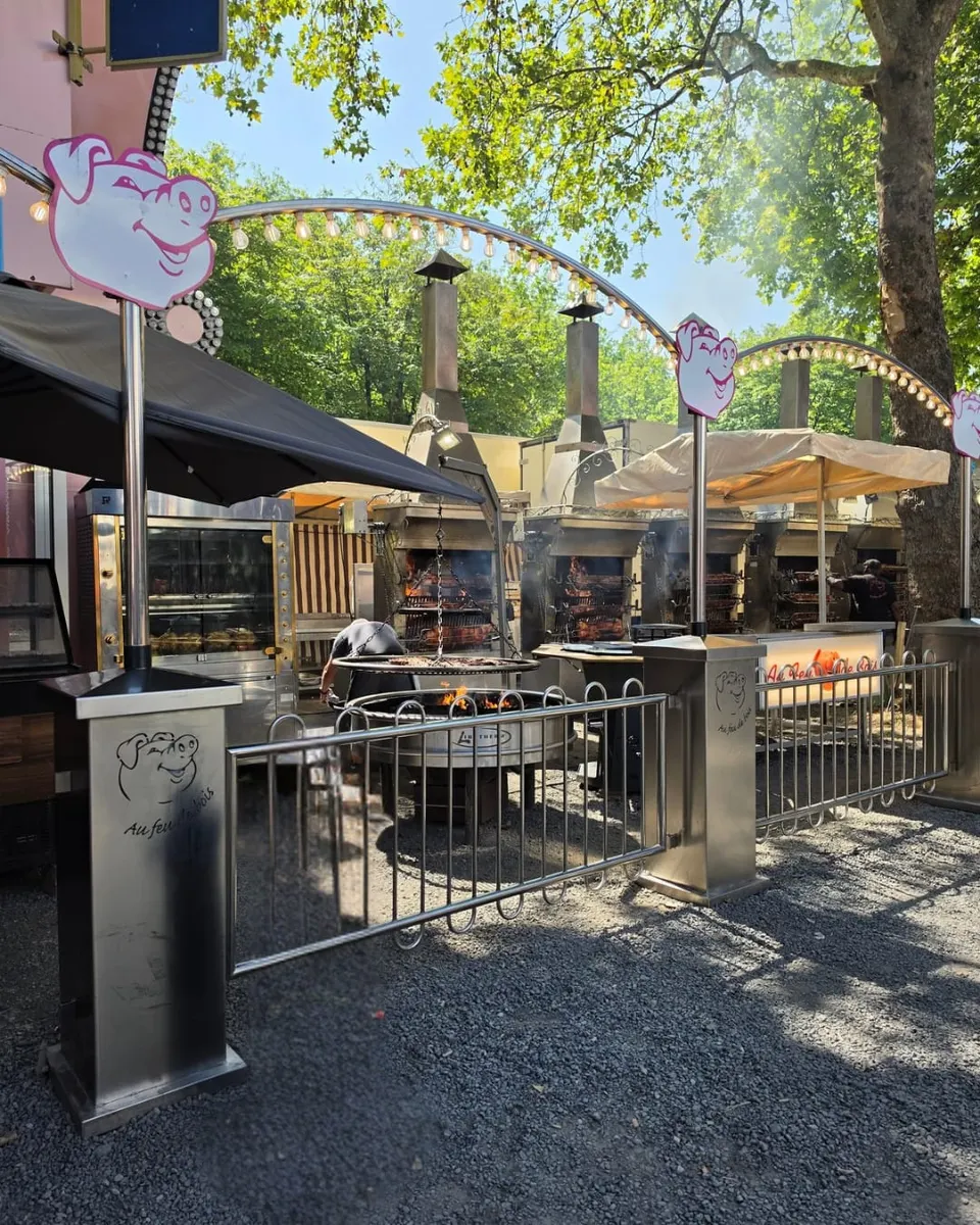 Espace de cuisson en plein air avec un grand brasero où des viandes grillent au-dessus des flammes, une rangée de rôtissoires et des arches lumineuses décorées de panneaux en forme de cochon.