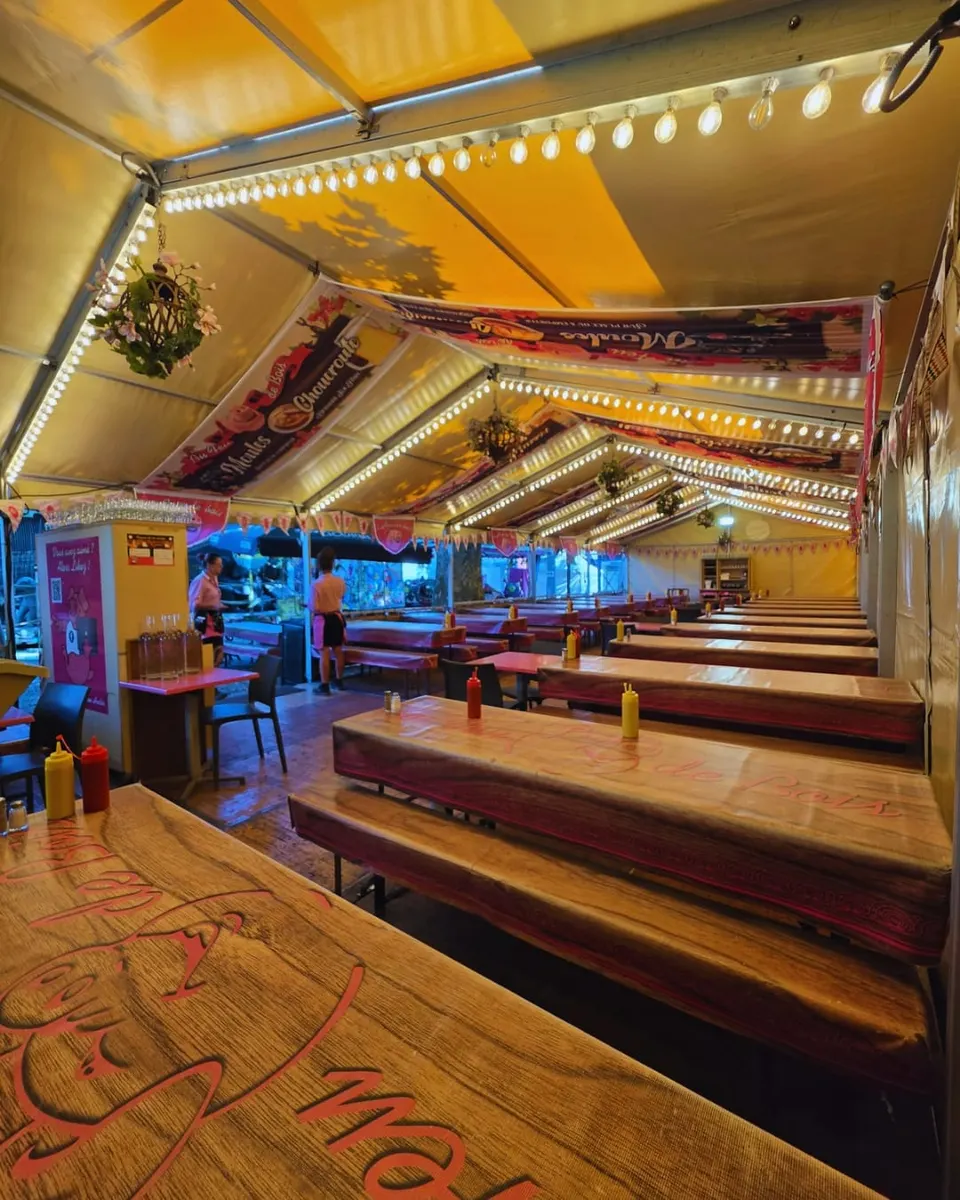 Salle sous chapiteau avec ses longues tablées en bois alignées sous des guirlandes d'ampoules, dressées et prêtes avant le service