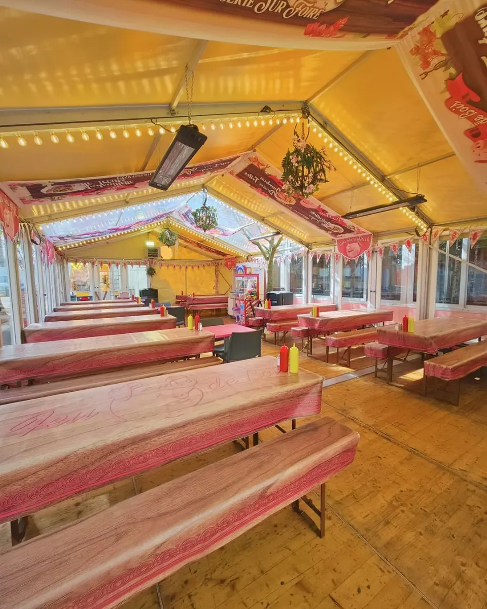 Salle sous chapiteau avec de longues tablées et bancs en bois rosé, des guirlandes d'ampoules et des fanions roses, avant l'arrivée des clients