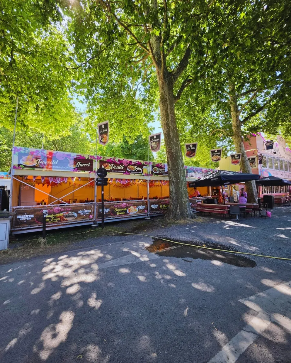 Vue d'ensemble du stand Au Feu de Bois installé sous les platanes d'une allée de fête foraine, avec sa terrasse et ses parasols le long de l'allée