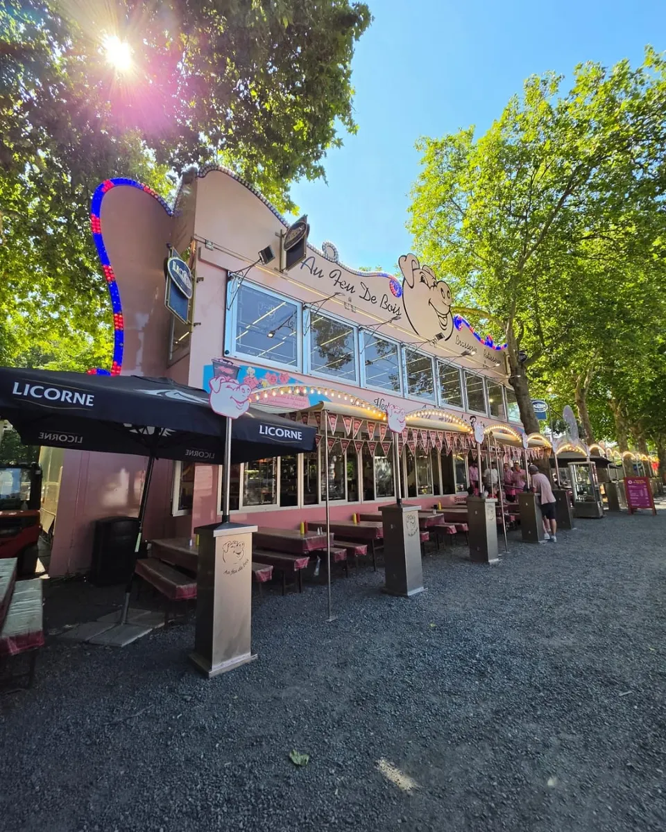 Vue de trois quarts de la brasserie foraine rose montrant la longue terrasse aux grandes tablées nappées de vichy rose sous des parasols, par une journée de plein soleil.