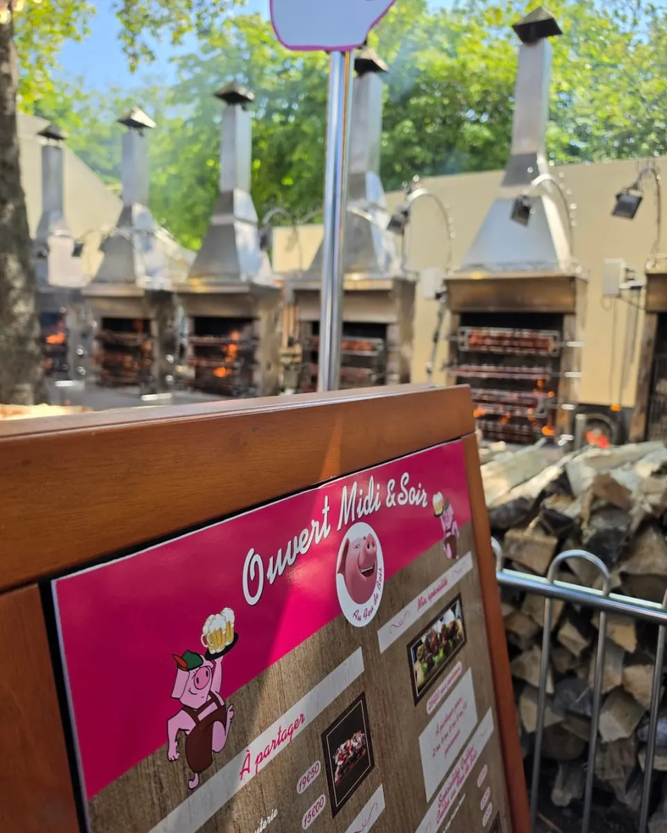 Panneau de menu en bois portant la mention Ouvert Midi et Soir et un cochon dessiné avec une chope de bière, devant les rôtissoires en train de cuire et une pile de bûches.