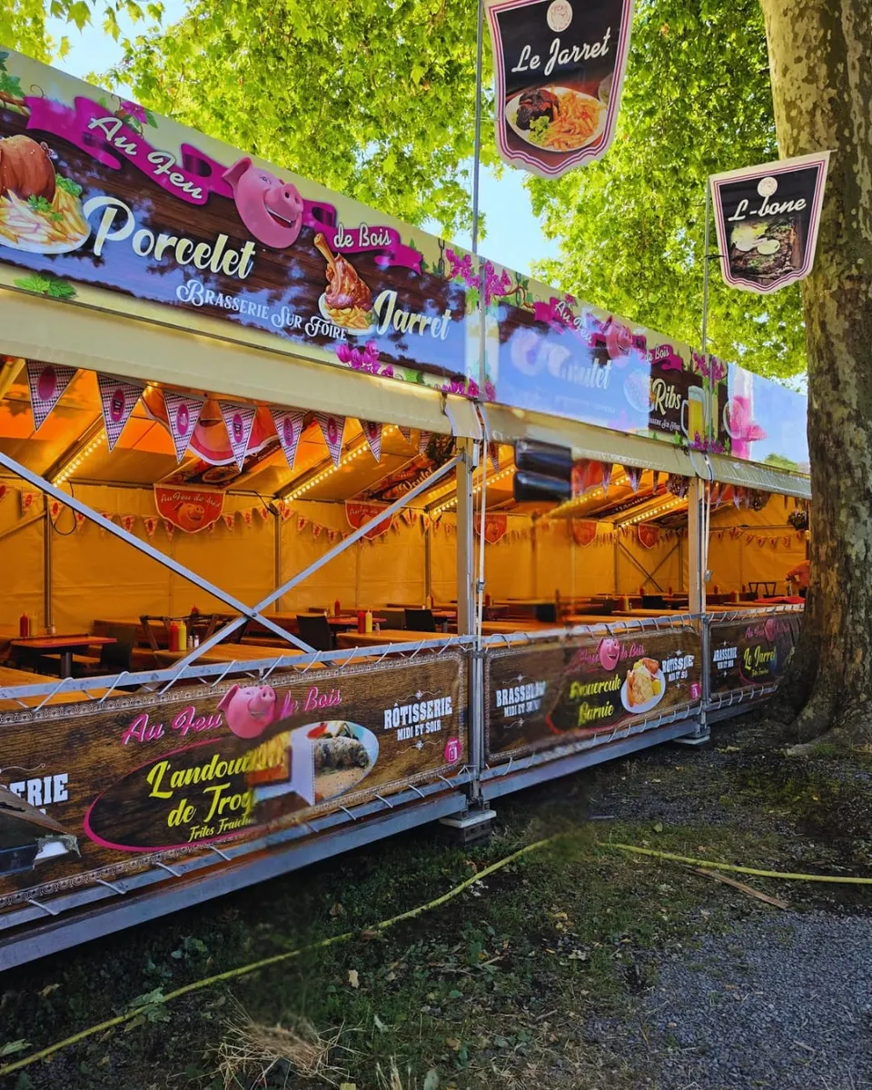 Remorque brasserie Au Feu de Bois installée sous les platanes, ses panneaux roses annonçant porcelet, jarret et andouillette de Troyes au-dessus des longues tablées en bois