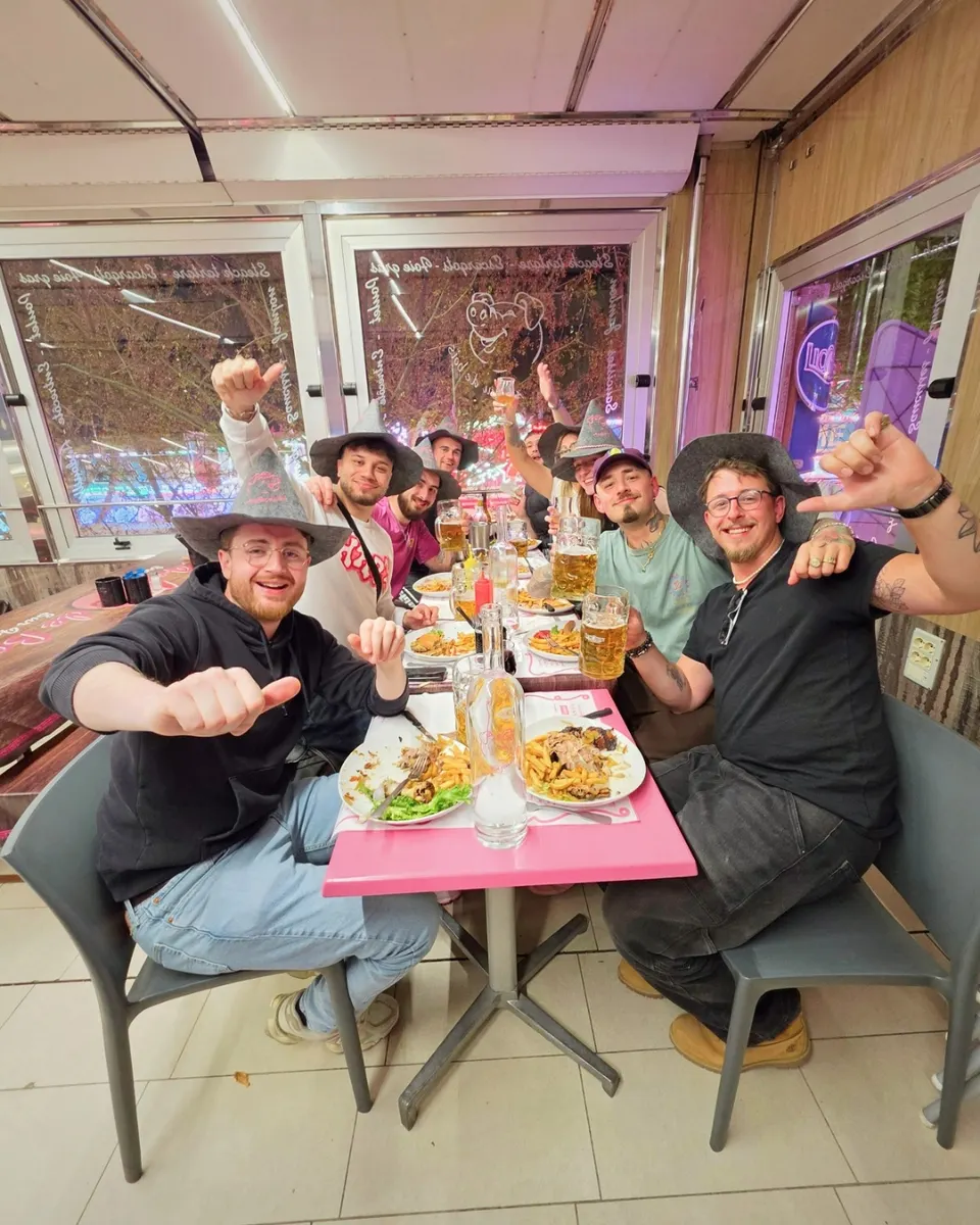 Bande d'amis en chapeaux de feutre autour d'une table rose, chopes levées et pouces tendus vers l'objectif