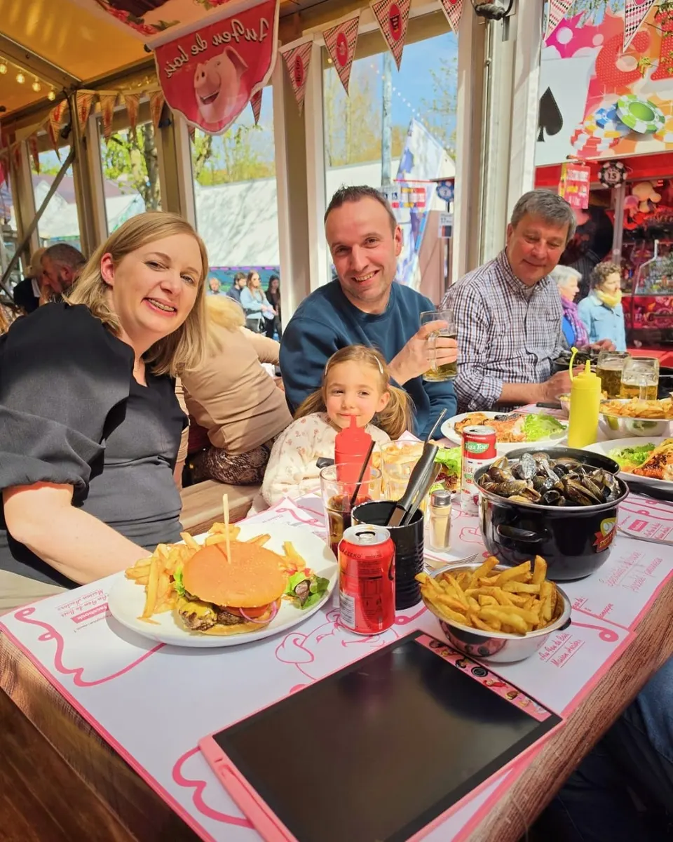 Un couple et leur petite fille attablés sourient devant un burger maison, une casserole de moules et un bol de frites.