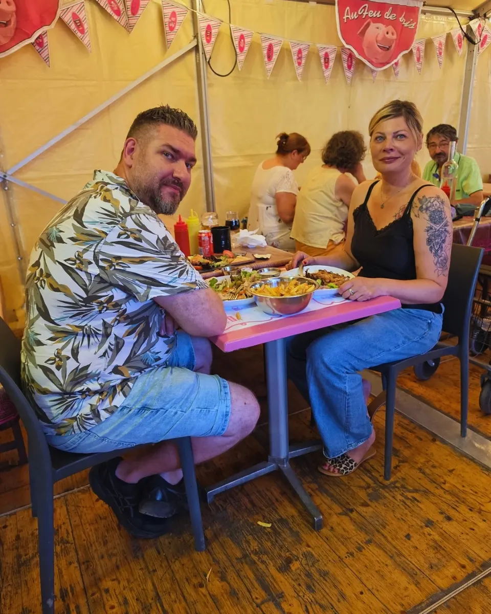 Couple attablé sous le chapiteau devant une assiette de viande et un saladier de frites sur une petite table rose