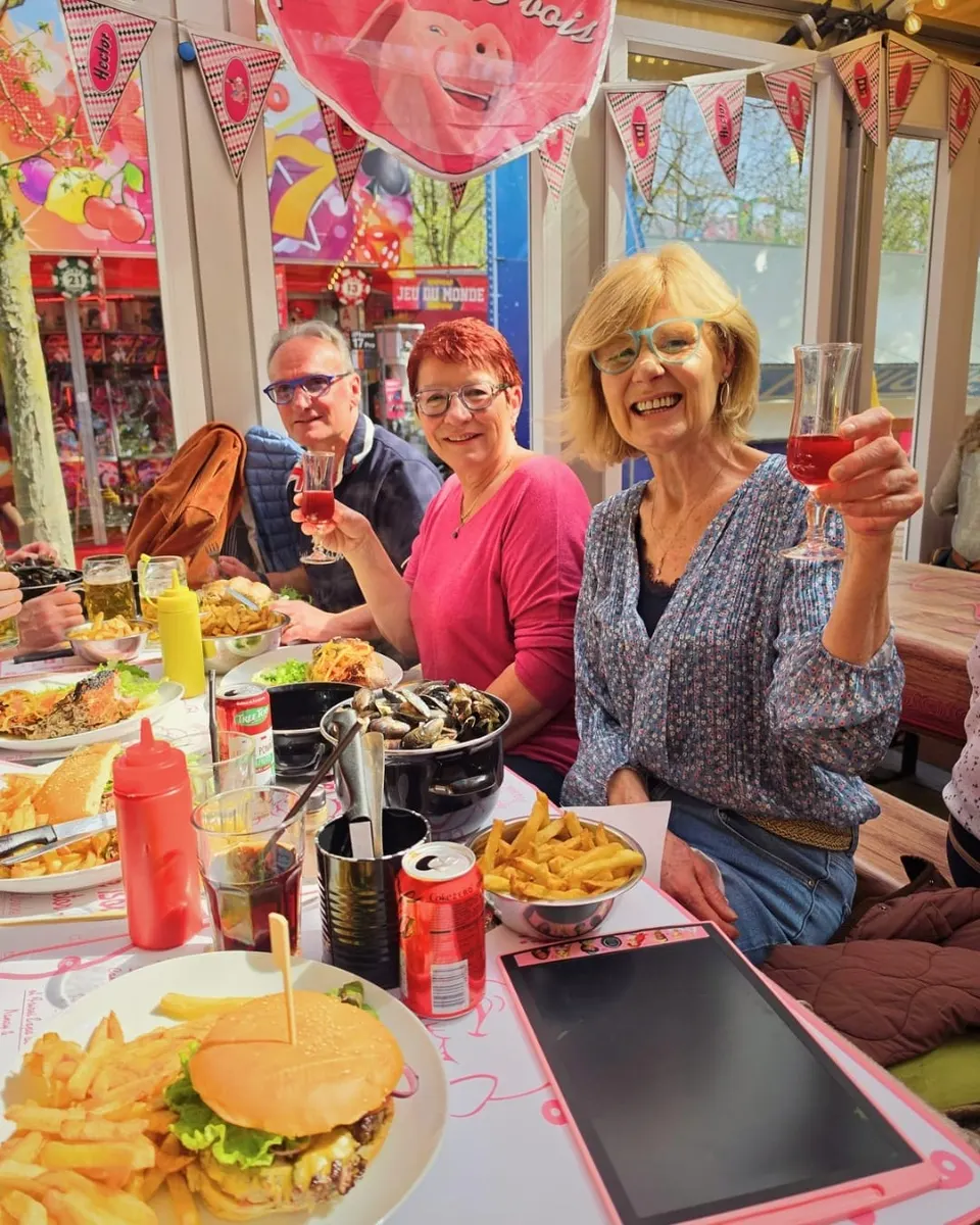 Deux clientes souriantes lèvent leur verre de kir au-dessus d'une table garnie de frites, de moules et d'un burger, sous les fanions roses de la brasserie.