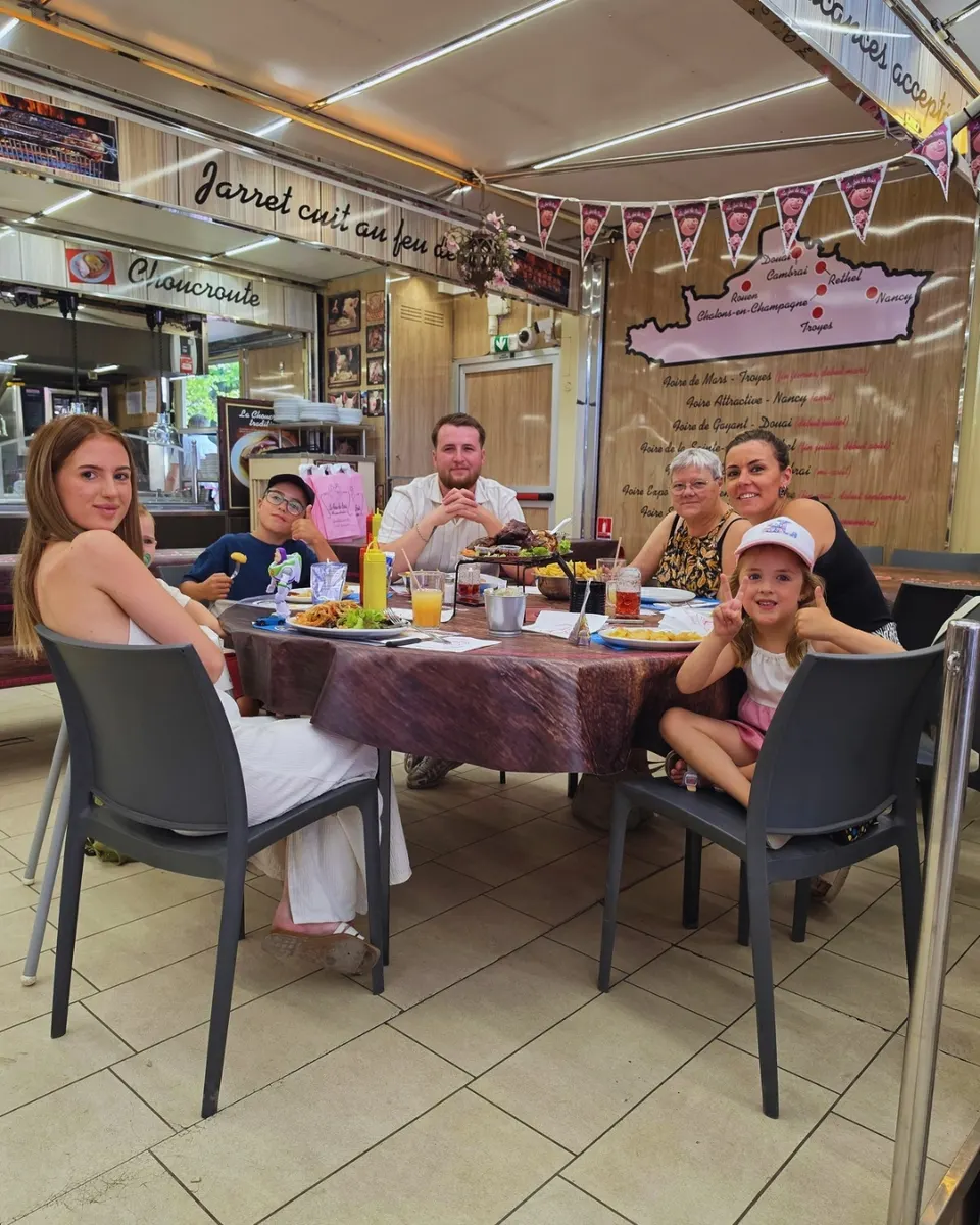 Une famille avec enfants attablée autour d'une table ronde en salle, devant le mur affichant la carte des foires et les spécialités de la maison.