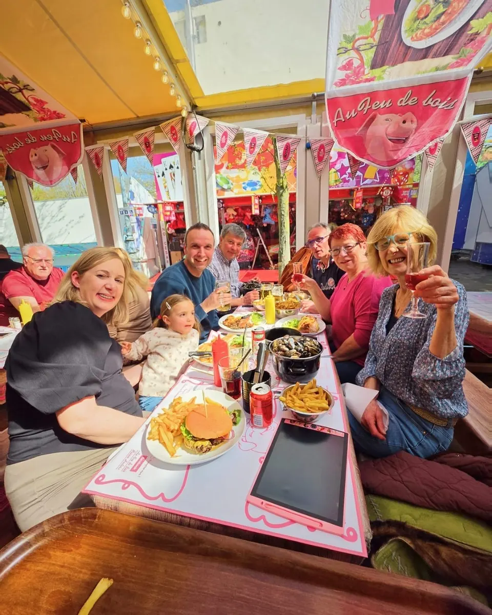 Une grande tablée familiale de huit personnes pose en souriant autour d'un repas, sous les bannières roses Au Feu de Bois et la mascotte cochon.