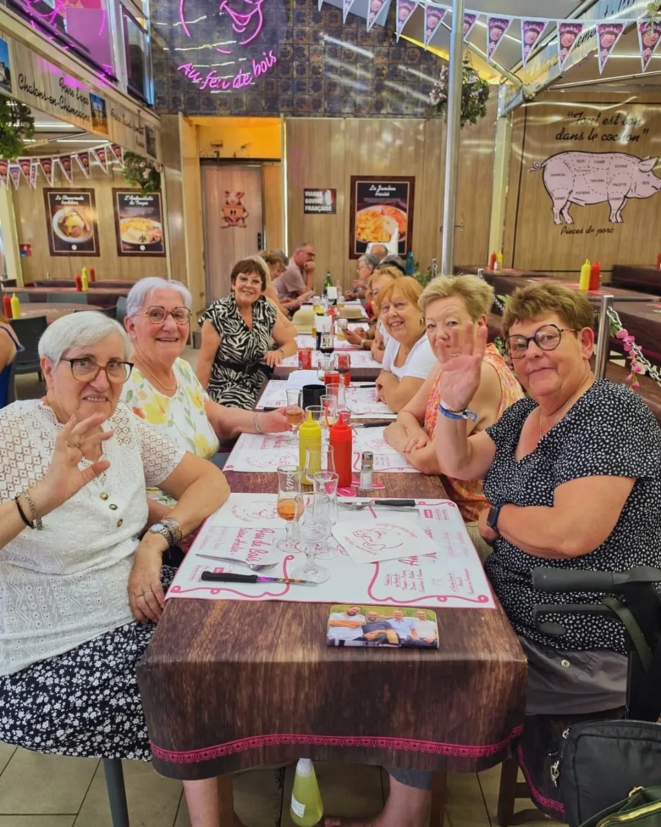 Un groupe de clientes souriantes salue l'objectif le long d'une grande tablée aux sets roses, sous l'enseigne néon Au Feu de Bois.