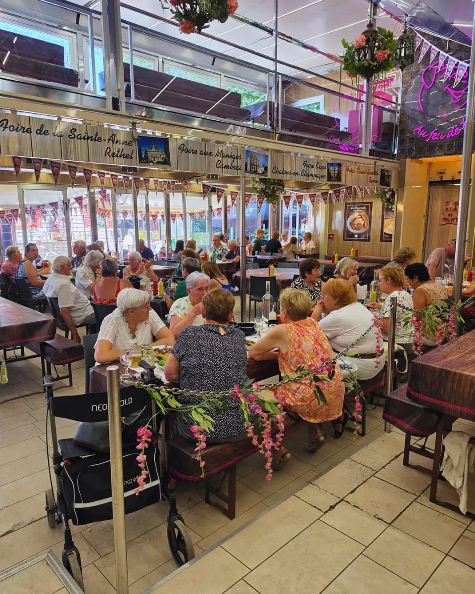 Salle comble de la brasserie foraine avec des clients installés aux grandes tablées en bois, sous les panneaux des foires et une enseigne néon rose