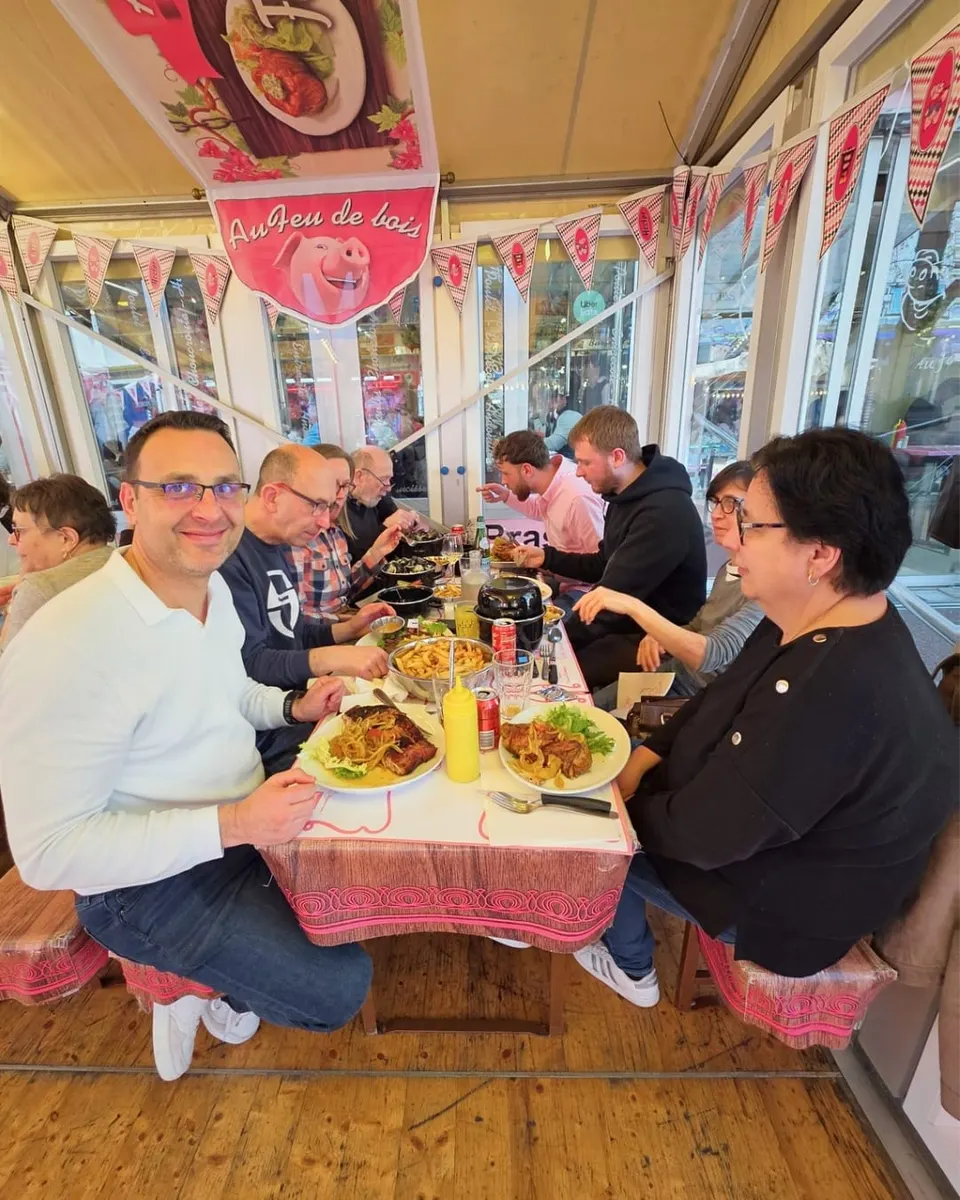 Un client souriant au premier plan d'une longue tablée pleine, devant une assiette de travers de porc grillé et un bol de frites.