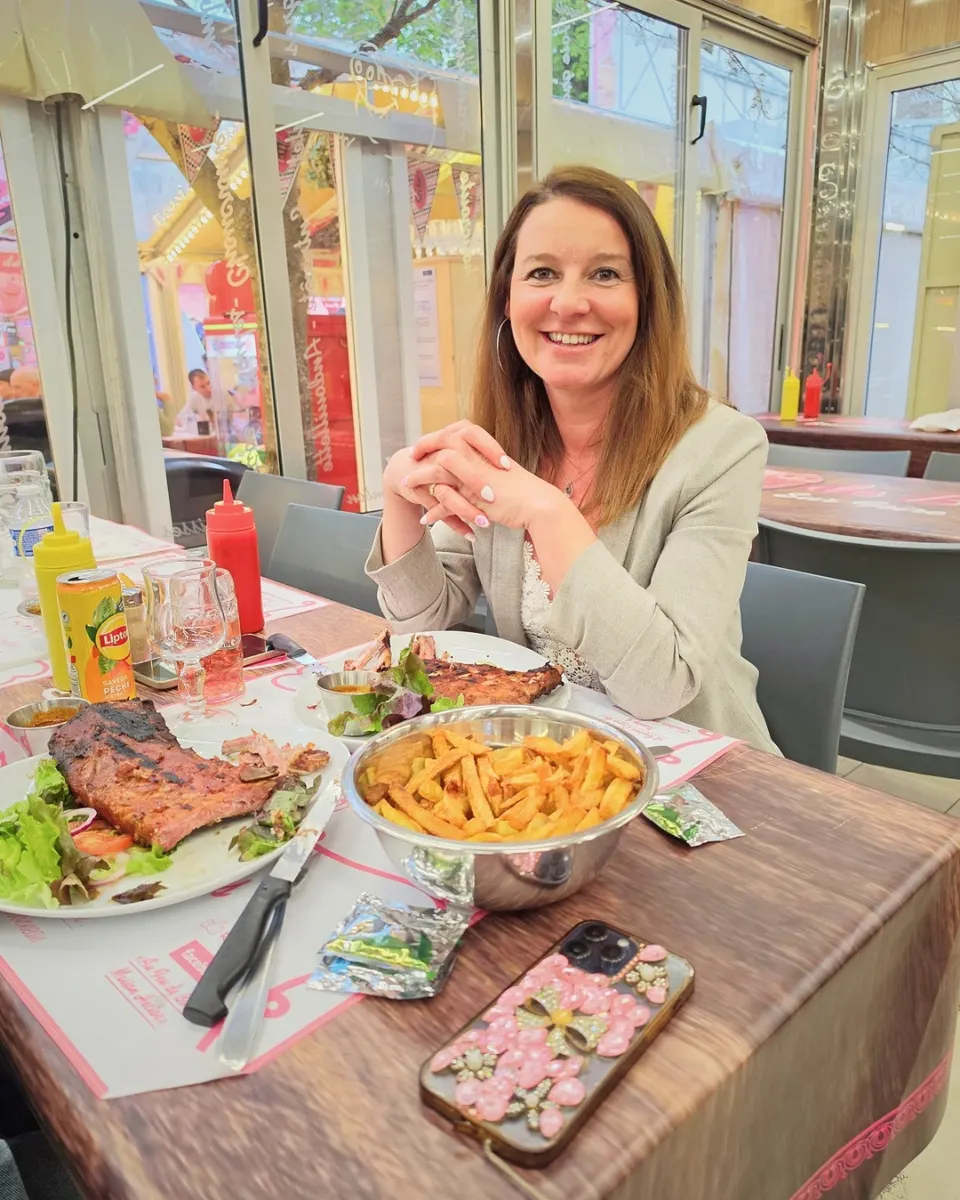 Une cliente souriante, mains jointes, pose à table devant deux assiettes de travers de porc grillés et un grand bol de frites.