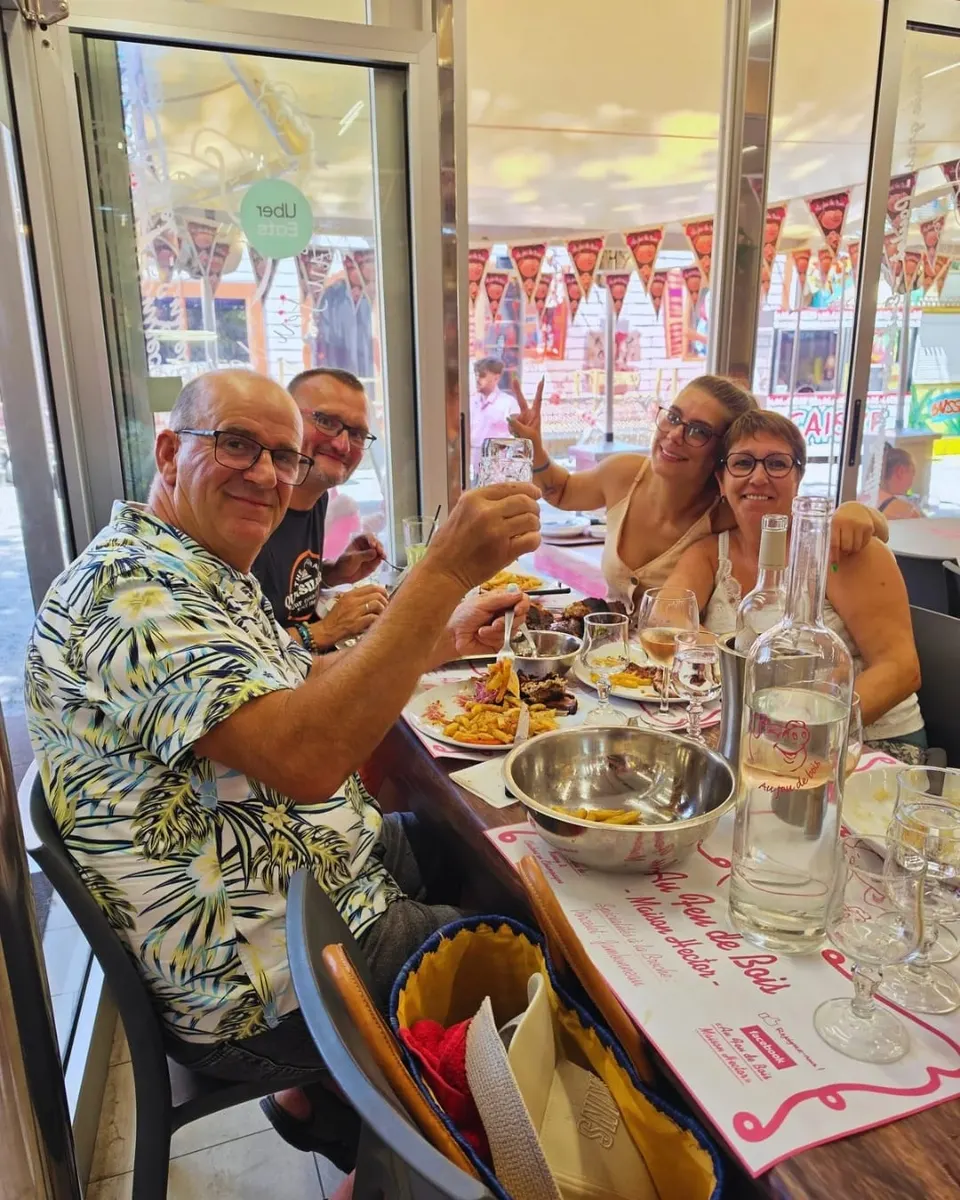 Quatre clients souriants lèvent leur verre autour d'une table dressée avec des sets roses Au Feu de Bois et des assiettes de viande grillée et de frites.