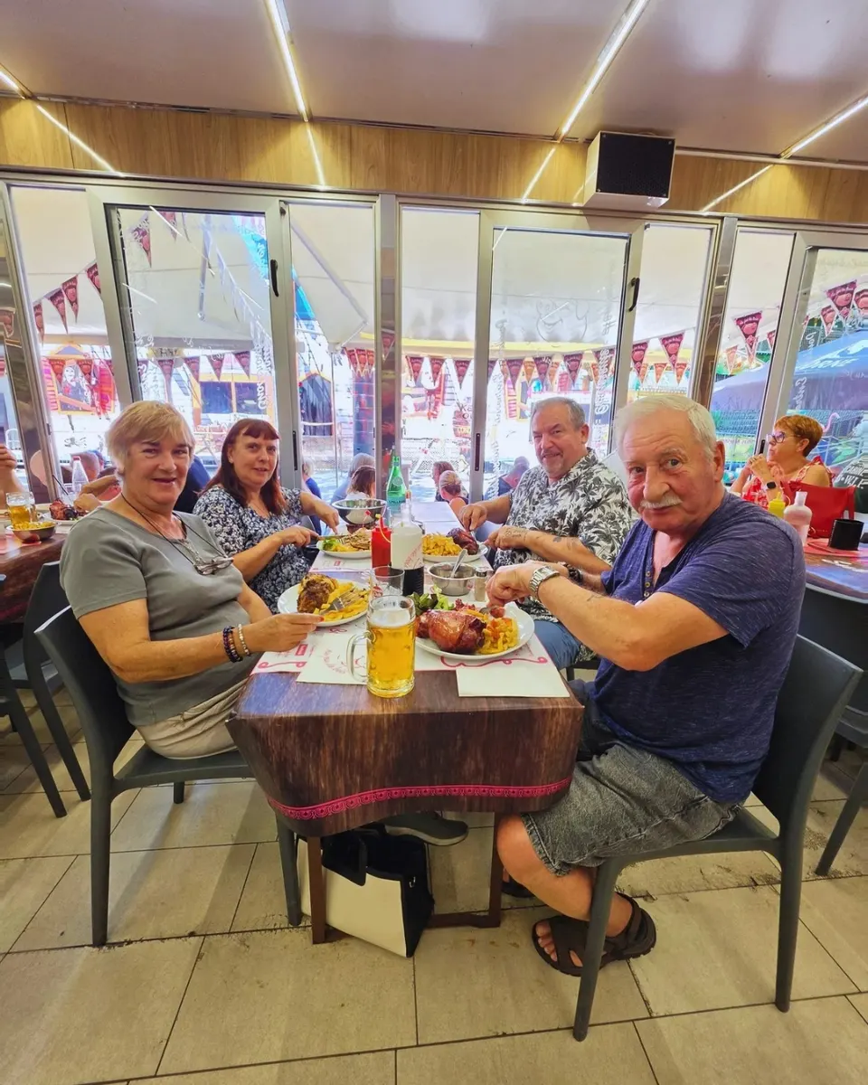 Quatre clients attablés en salle sourient devant des assiettes de jarret et de frites, avec une chope de bière au centre de la table.