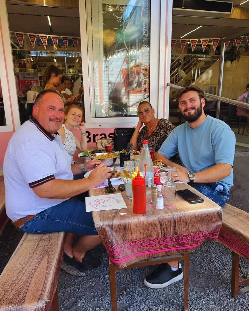 Quatre clients souriants attablés en terrasse sur le gravier devant la façade rose de la brasserie, avec un set de table à la mascotte cochon.