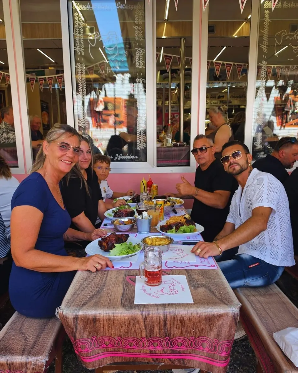 Six clients attablés en terrasse posent devant des assiettes de jarret grillé et de frites, devant la devanture rose vitrée du restaurant.