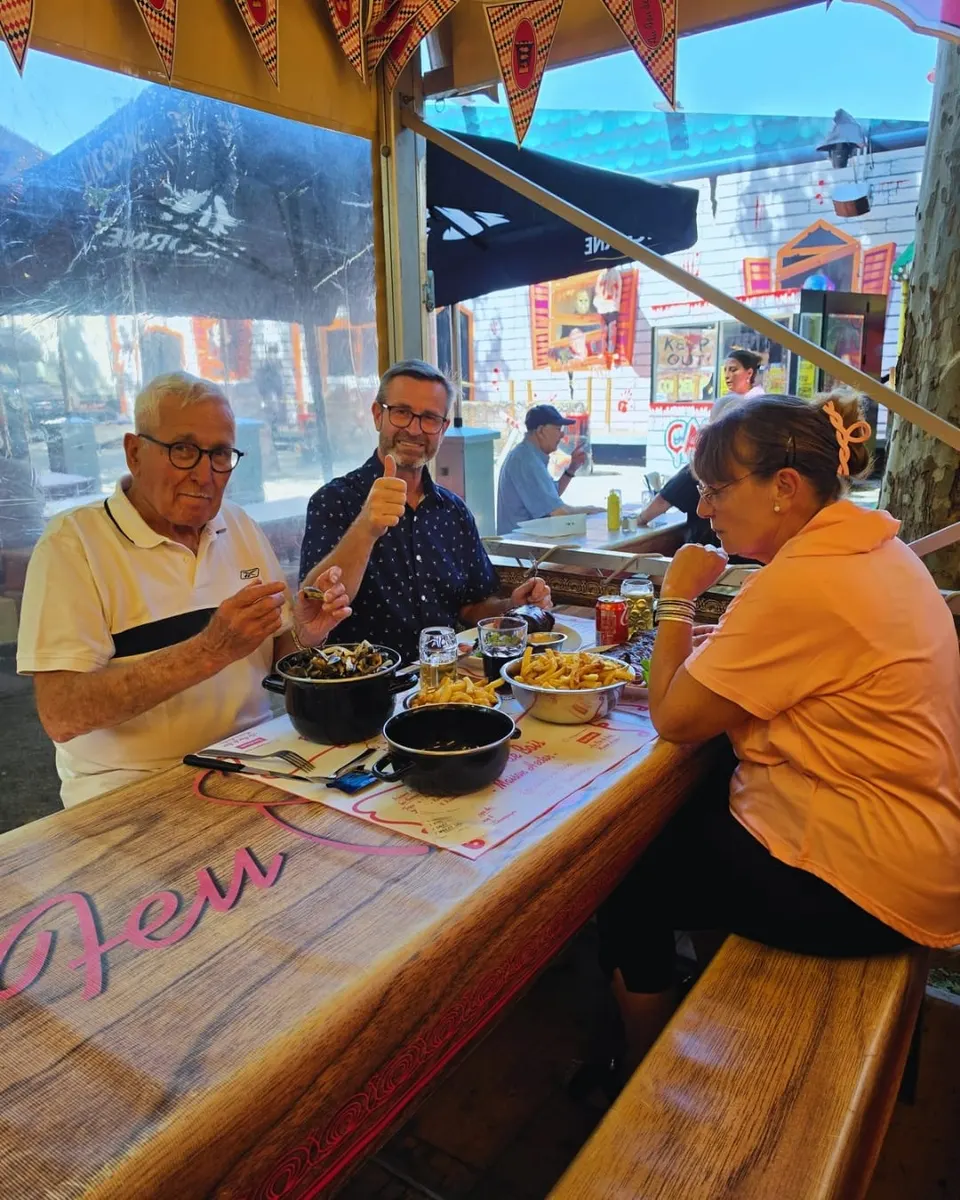 Trois clients attablés devant des marmites de moules et des frites, l'un d'eux levant le pouce vers l'objectif, sur une table au lettrage rose de la maison