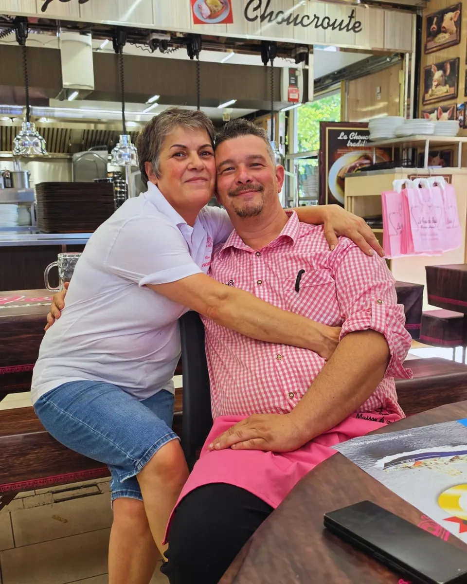 Les deux patrons souriants joue contre joue dans leur brasserie, elle en polo blanc, lui en chemise vichy rose.
