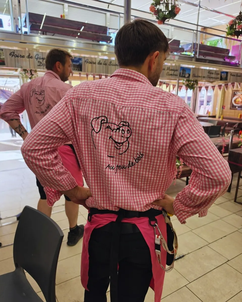 Deux membres de l'équipe de dos en chemise vichy rose brodée du cochon souriant, dans la halle de la foire.