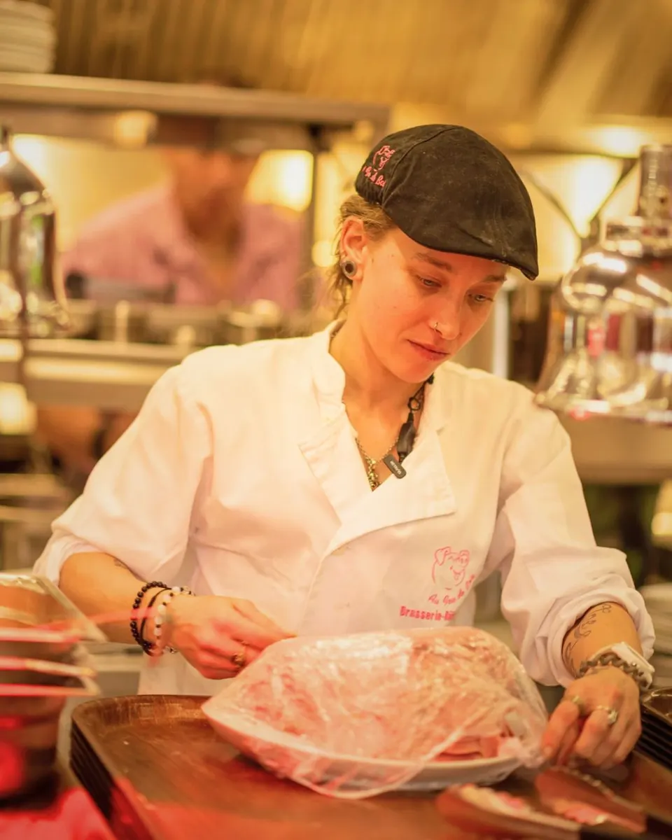 Une cuisinière en veste blanche et casquette noire prépare une grande pièce de viande sur un plateau en cuisine.