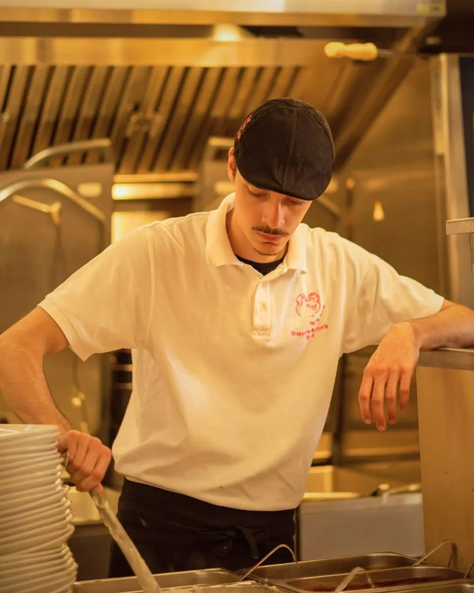 Un jeune membre de l'équipe en polo blanc et casquette noire travaille au passe de la cuisine, à côté d'une haute pile d'assiettes.