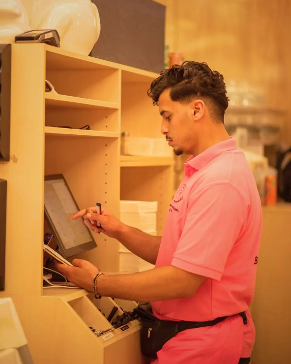 Un jeune serveur en polo rose enregistre une commande sur l'écran tactile de la caisse, son carnet à la main.