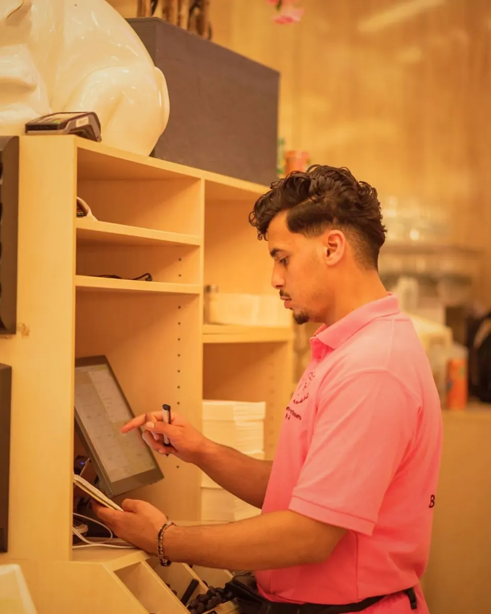 Jeune serveur en polo rose saisissant une commande sur un écran de caisse tactile, carnet à la main