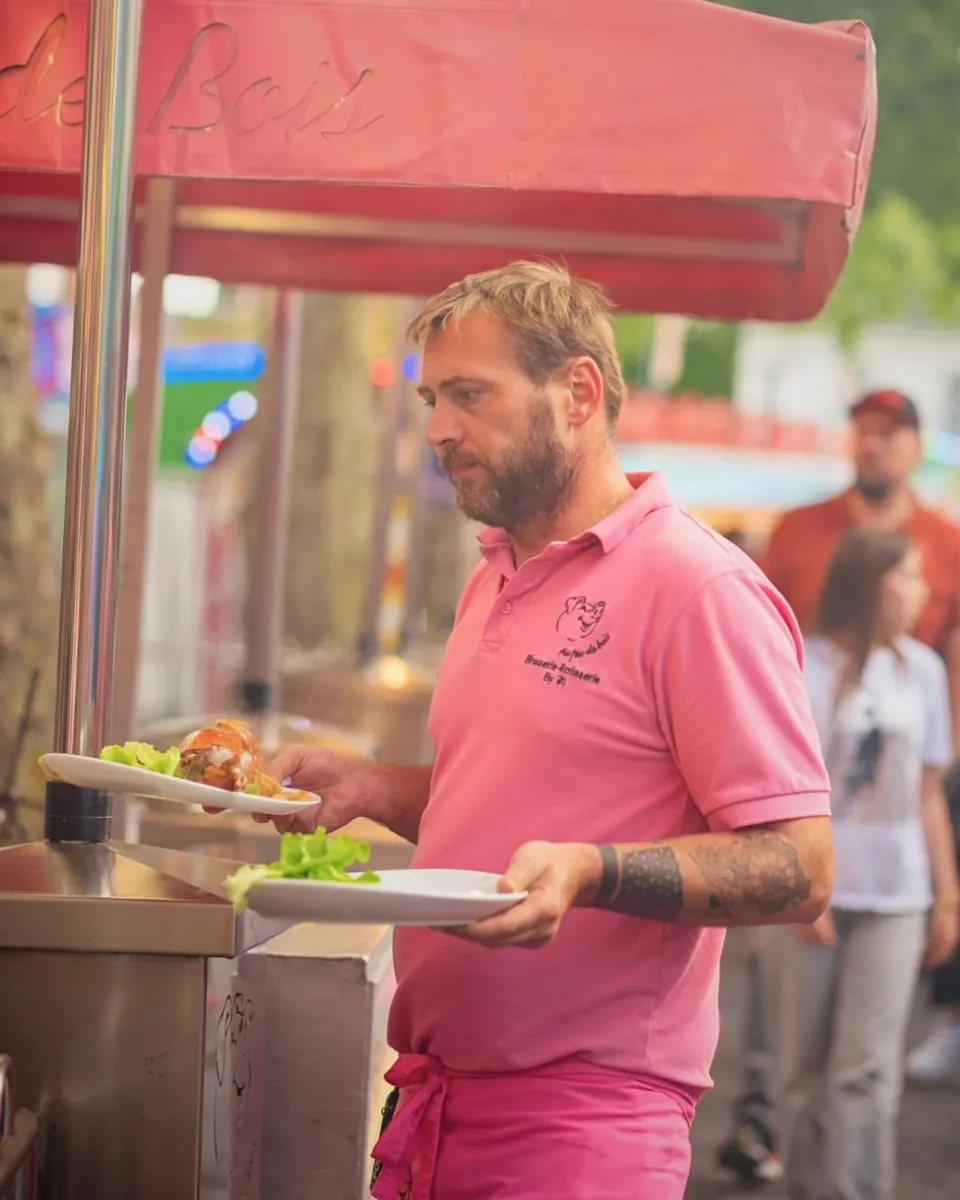 Un serveur en polo rose dresse deux assiettes de viande grillée et de salade sous l'auvent rose du stand, dans l'allée de la fête foraine.