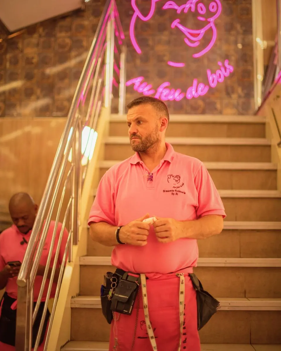 Homme en polo rose et tablier debout dans l'escalier sous l'enseigne néon rose représentant un cochon et le nom Au feu de bois