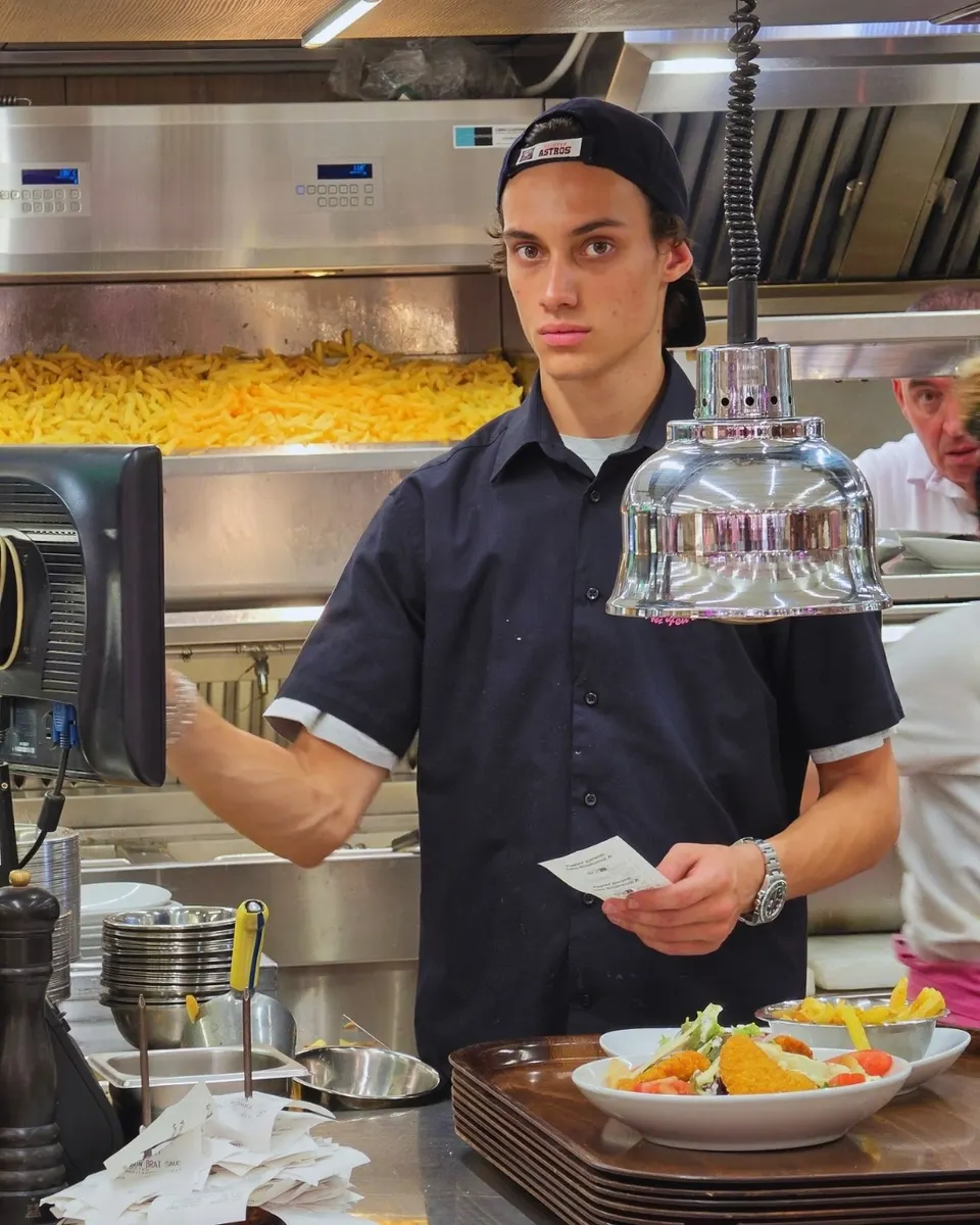 Jeune membre de l'équipe au poste d'envoi de la cuisine, bon de commande en main devant le bac de frites dorées.