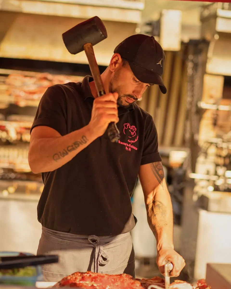 Un rôtisseur en polo noir lève un maillet pour découper un cochon grillé, devant la rôtissoire où cuisent d'autres pièces.