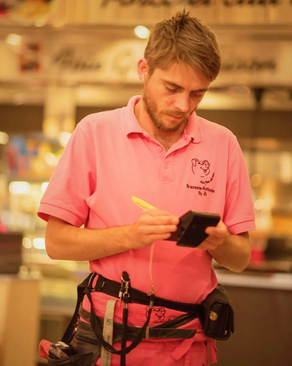 Un serveur en polo rose note une commande sur un terminal portable, un stylo jaune à la main, dans la salle du restaurant.