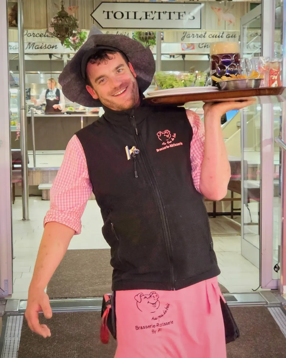Serveur souriant avec chapeau et chemise vichy rose portant à l'épaule un plateau de plats et de bières à l'entrée du restaurant