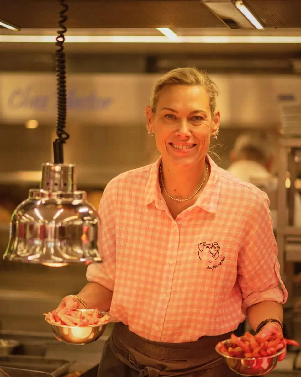 Une serveuse en chemise à carreaux roses sourit en cuisine, une coupelle de frites dans chaque main, sous une lampe chauffante.