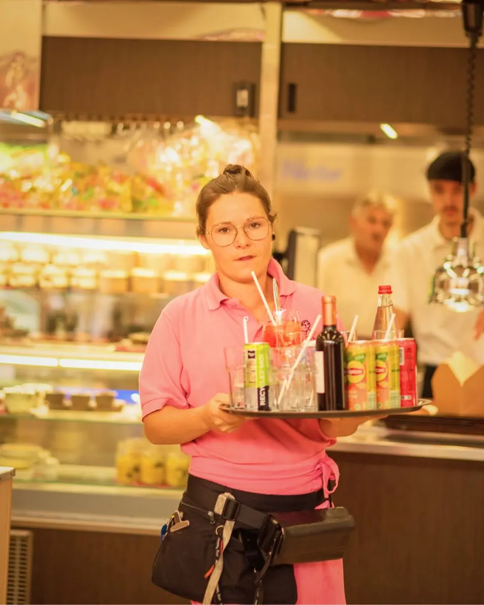 Serveuse à lunettes en polo rose portant un plateau rempli de boissons devant la vitrine du comptoir