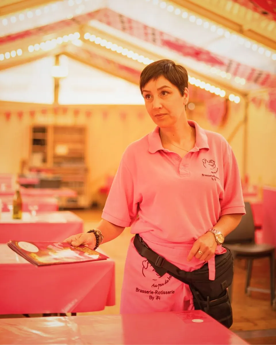 Serveuse en polo rose de la brasserie posant une carte de menu sur une table nappée de rose sous le chapiteau illuminé