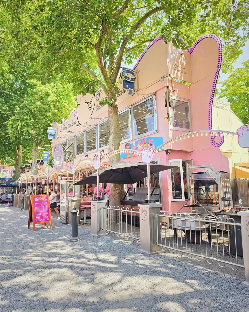 La façade rose du stand sous les platanes : fronton à ampoules, guirlandes, terrasse et rôtissoires en plein air.