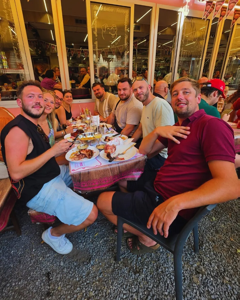 Groupe d'amis souriants attablés en terrasse devant grillades et frites, vitres roses au cochon du logo.