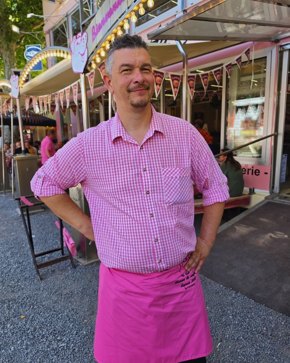 Le patron de la maison, mains sur les hanches, en chemise vichy et tablier fuchsia devant sa brasserie illuminée