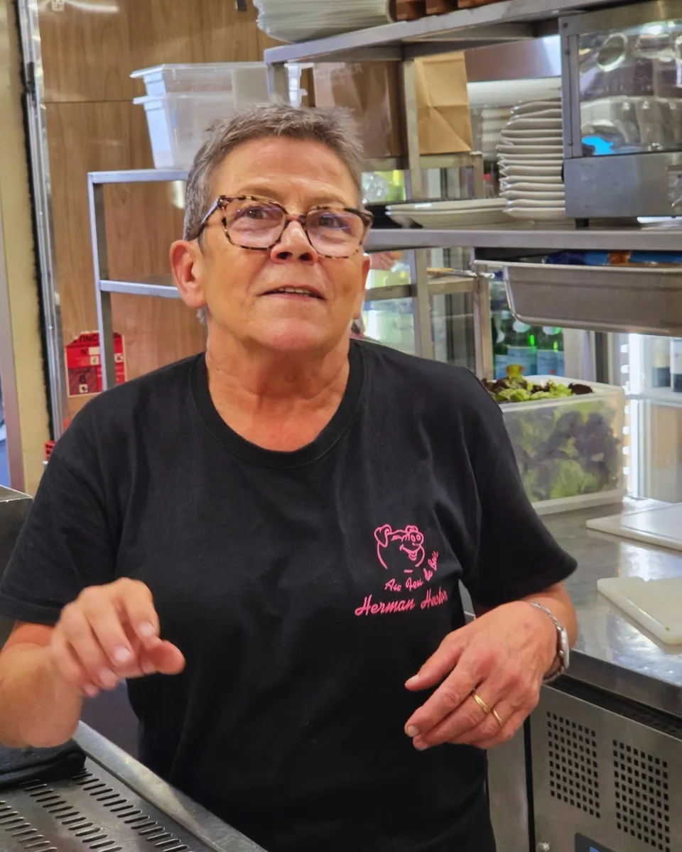 Membre de l'équipe souriante derrière le comptoir de la cuisine, en t-shirt noir au logo cochon rose Au Feu de Bois.